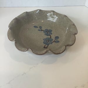 May include: A ceramic bowl with a scalloped edge and a speckled, muted green finish. The bowl features a floral design in blue. The rim has a brown trim.