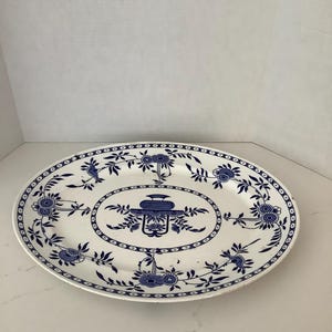 May include: An oval ceramic platter featuring a white base with a blue transferware pattern. The design includes a central urn motif surrounded by an oval border, with floral and leaf motifs extending toward the scalloped rim edge.