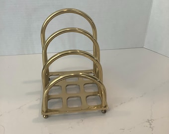 Puede incluir: Un tarjetero de latón pulido con cuatro divisores arqueados de tamaño gradual conectados a una base cuadrada con patrón de rejilla. La estructura se apoya sobre cuatro pequeños pies esféricos en cada esquina, proporcionando una base estable.