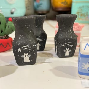 May include: Three black ceramic vases with white cat and star designs. The vases are speckled with white paint and have a matte finish. The vases are arranged on a white surface.