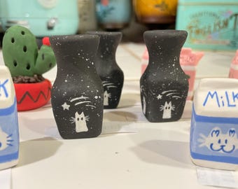 May include: Three black ceramic vases with white cat and star designs. The vases are speckled with white paint and have a matte finish. The vases are arranged on a white surface.