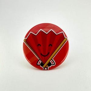May include: A round, red button featuring a cartoon fan design. The fan is red with white accents and a smiling face. The fan is held by two golden-yellow sticks. The button is a fun accessory.