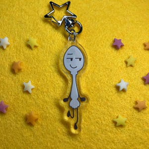 May include: A clear acrylic keyring featuring a cartoon spoon character with a smug expression. The keyring has a silver star-shaped clasp and ring. The background is yellow, with scattered star-shaped sprinkles in various colours.