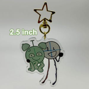 May include: A 2.5 inch clear acrylic keychain featuring a green cartoon character with a red and blue stick figure character. The keychain has a gold star-shaped swivel clasp.