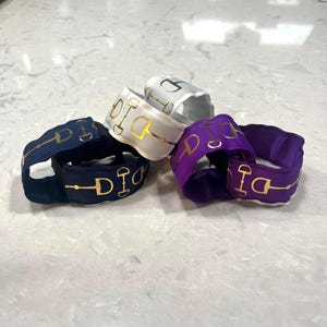 Peut inclure: Plusieurs bracelets de poignet sur le thème équestre, de couleurs marine, blanc, argent et violet. Chaque bracelet présente un motif de mors doré. Les bracelets sont faits d'un tissu côtelé et sont empilés sur une surface claire.