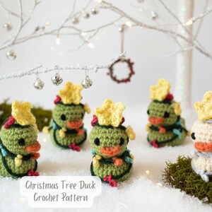 May include: A collection of crocheted Christmas tree ducks. The ducks are primarily green with yellow star-shaped hats, red accents, and orange beaks. They are arranged on a snowy surface with decorative elements in the background.