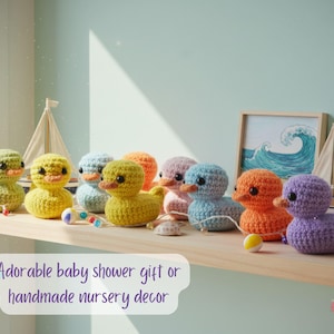 Pu&ograve; includere: Una collezione di anatroccoli all'uncinetto colorati in varie tonalit&agrave; di verde, giallo, blu, arancione, rosa e viola. Le anatre fatte a mano sono esposte su una mensola di legno con una piccola barca a vela e un'immagine incorniciata di un'onda.