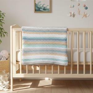 May include: A handmade baby blanket in a crib. The blanket features horizontal stripes in shades of white, blue, gray, and tan. The crib is made of light-colored wood. A mobile with sea-themed decorations hangs above the crib.