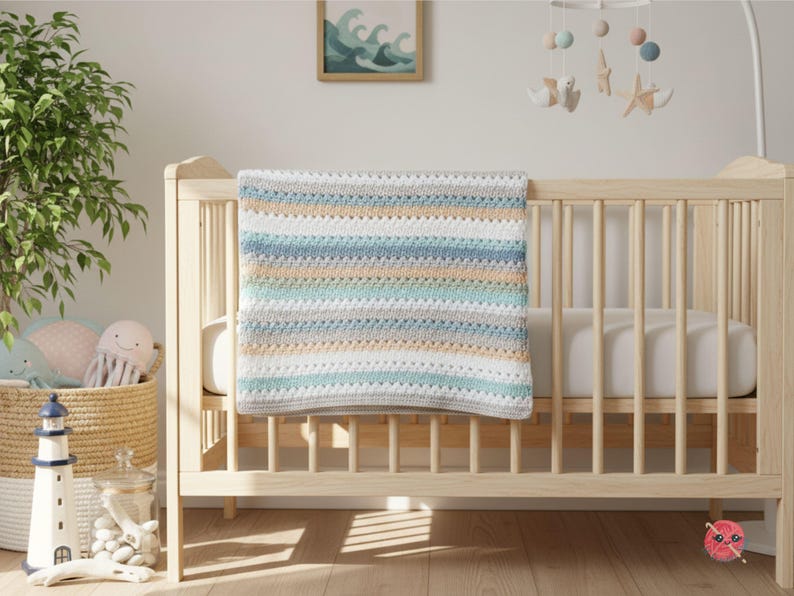 May include: A handmade baby blanket in a crib. The blanket features horizontal stripes in shades of white, blue, gray, and tan. The crib is made of light-colored wood. A mobile with sea-themed decorations hangs above the crib.