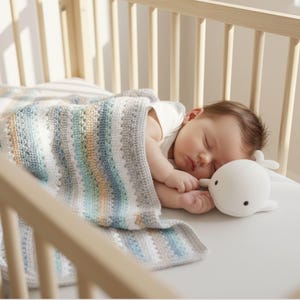 May include: A sleeping baby in a wooden crib, covered with a striped, multi-colored blanket. A white whale-shaped plush toy rests beside the baby. The blanket features stripes of blue, gray, and tan.