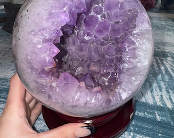 May include: A large, spherical amethyst geode, showcasing a deep purple crystalline interior. The geode is set on a dark wooden stand, with a polished exterior. Other geodes are visible in the background.