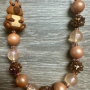 May include: A necklace featuring a brown capybara-shaped bead, interspersed with round beads in shades of tan, peach, and translucent pink, along with dark brown textured glitter beads, all strung on a white cord with small metallic spacers.