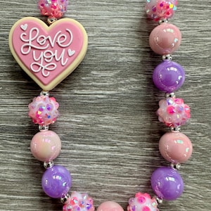 Puede incluir: Un collar de cuentas rosas y moradas con un colgante en forma de corazón. El colgante tiene la inscripción "Love You". El collar presenta cuentas redondas y cuentas brillantes.