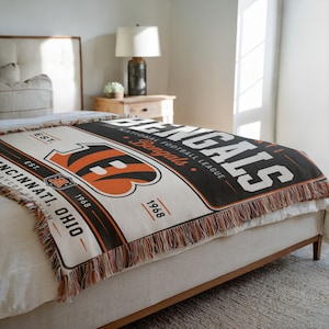 Peut inclure: Un plaid tissé arborant le logo et le texte des Cincinnati Bengals. Le motif comprend le nom de l'équipe, l'année 1968 et la ville de Cincinnati, Ohio, dans une palette de couleurs noir, orange et crème avec des bords à franges décoratives.