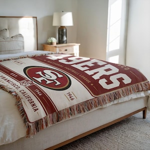 May include: A woven throw blanket featuring the San Francisco 49ers logo, the text 49ERS, NATIONAL FOOTBALL LEAGUE, EST. 1946, and SAN FRANCISCO, CALIFORNIA. The design includes a large central team emblem in red, gold, and white with fringe edges.