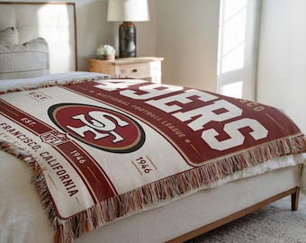 May include: A woven throw blanket featuring the San Francisco 49ers logo, the text 49ERS, NATIONAL FOOTBALL LEAGUE, EST. 1946, and SAN FRANCISCO, CALIFORNIA. The design includes a large central team emblem in red, gold, and white with fringe edges.