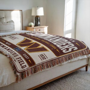 May include: A woven throw blanket featuring the Washington Commanders logo and text, including 'EST. 1932', 'NFC EAST', and 'COMMANDERS FIELD'. The design uses a burgundy, gold, and cream colour palette with decorative fringe along the edges.