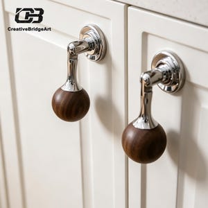 May include: Two cabinet knobs featuring a spherical dark brown wood handle attached to a polished chrome metal base. The hardware is mounted on white cabinet doors. The image includes the logo text CreativeBridgeArt in the top left corner.
