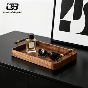 May include: A rectangular wooden tray with a natural brown grain finish, featuring two horizontal wooden handles secured by gold-toned metal brackets. Inside the tray sits a clear glass perfume bottle with a black cap and black sunglasses.