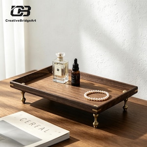 May include: A rectangular dark wood tray with four brass-toned metal feet and brass corner accents. On the tray sits a clear glass perfume bottle, a small dark amber glass dropper bottle, and a single strand of white pearls arranged in a circle.