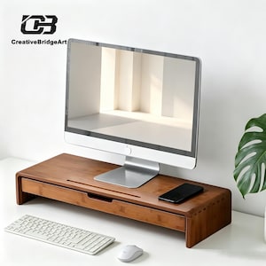 May include: A rectangular bamboo monitor stand with a built-in storage drawer, featuring a smooth wood grain finish. The stand supports a silver computer monitor, with a black smartphone resting on the surface and a white keyboard and mouse nearby.