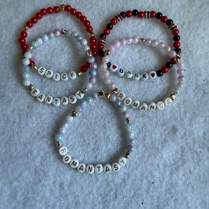 May include: Five beaded bracelets in red, black, and iridescent colors. The bracelets feature gold accents and letter beads spelling out "SPICY", "FANTASY", "DARK", "ROMANCE", and "ROMANTASY".