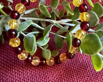 May include: A beaded bracelet featuring alternating amber and dark red round beads. The bracelet is displayed on a burgundy surface with green leafy sprigs.