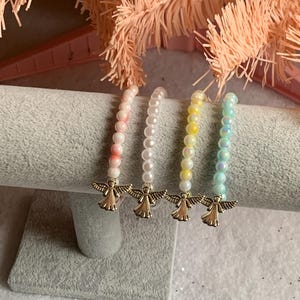 May include: Four beaded bracelets in shades of pink, white, yellow, and iridescent blue-green. Each bracelet features a gold-colored angel charm. The bracelets are displayed on a gray, textured surface.