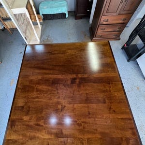 May include: A large, square, wooden table top with a rich brown stain. The table top is made of multiple pieces of wood that have been joined together to create a unique pattern.