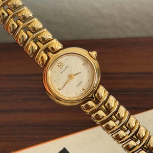 Könnte beinhalten: Eine goldfarbene Citizen Sylph Armbanduhr mit cremefarbenem Zifferblatt und goldenen Zeigern. Die Uhr hat ein goldenes Armband mit einem einzigartigen Gliederdesign. Das Zifferblatt zeigt den Markennamen und die Zahl XII.