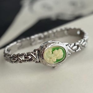 May include: Silver-toned bracelet featuring an oval cameo with a light green background and a white profile. The bracelet has an intricate, decorative link design. The cameo is set in a detailed frame. The bracelet is on a white surface.