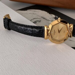 May include: A gold-tone wristwatch with a black leather band. The watch face is octagonal and features a gold-tone dial with diamond hour markers and a date window. The watch is made by Pierre Cardin.