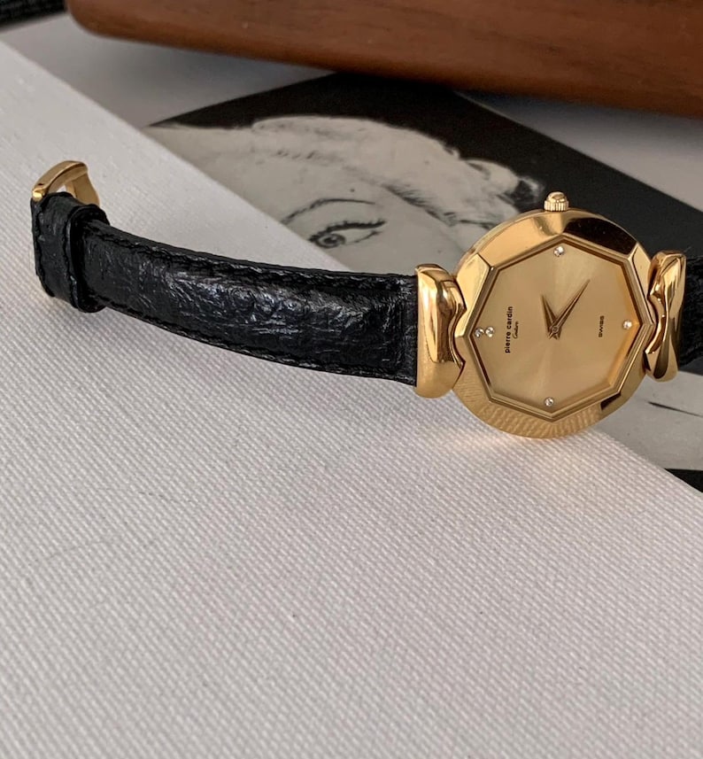 May include: A gold-tone wristwatch with a black leather band. The watch face is octagonal and features a gold-tone dial with diamond hour markers and a date window. The watch is made by Pierre Cardin.