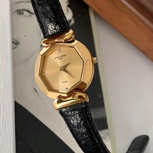 May include: A gold-tone Pierre Cardin watch with a black leather band. The watch face is octagonal and has a gold-tone bezel with small diamonds around the edge. The watch face is gold-tone and has black hands and markers.