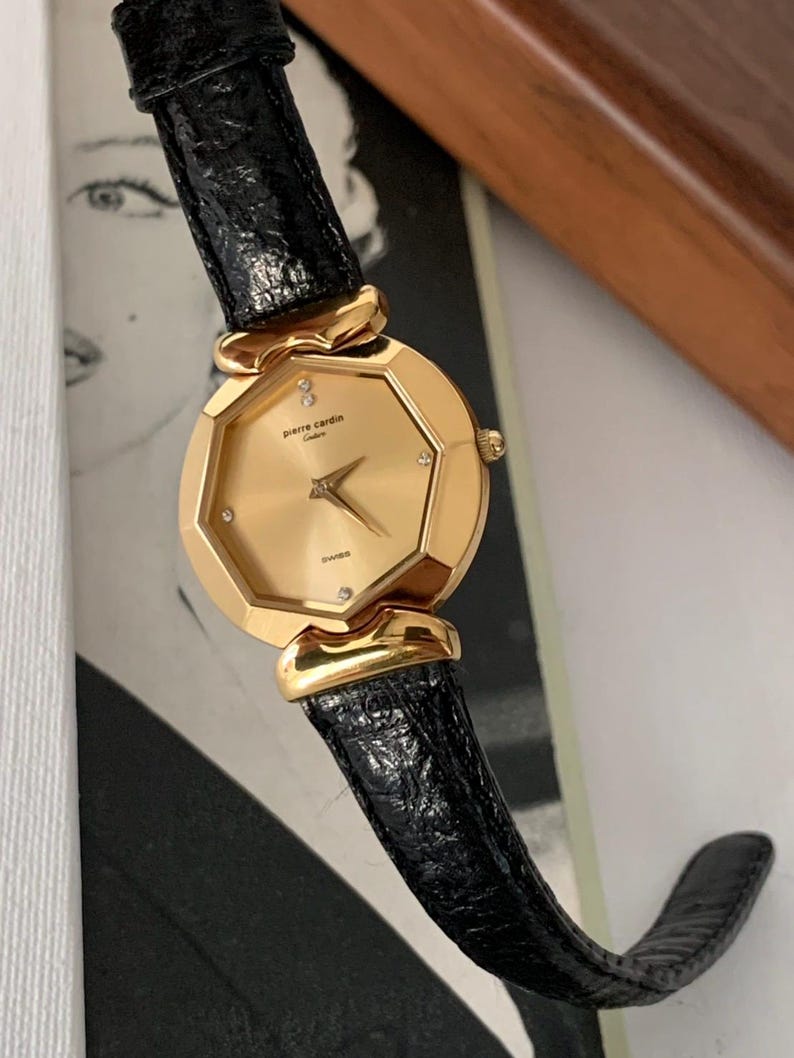 May include: A gold-tone Pierre Cardin watch with a black leather band. The watch face is octagonal and has a gold-tone bezel with small diamonds around the edge. The watch face is gold-tone and has black hands and markers.