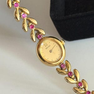May include: A gold-tone Seiko watch with a round face, gold dial, and Roman numeral at the twelve o'clock position. The bracelet features a repeating leaf-shaped link design accented with small, round, vibrant pink stones set between each leaf.