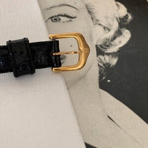 May include: A gold-tone watch strap buckle with a black leather strap. The buckle has a simple design with a smooth finish.