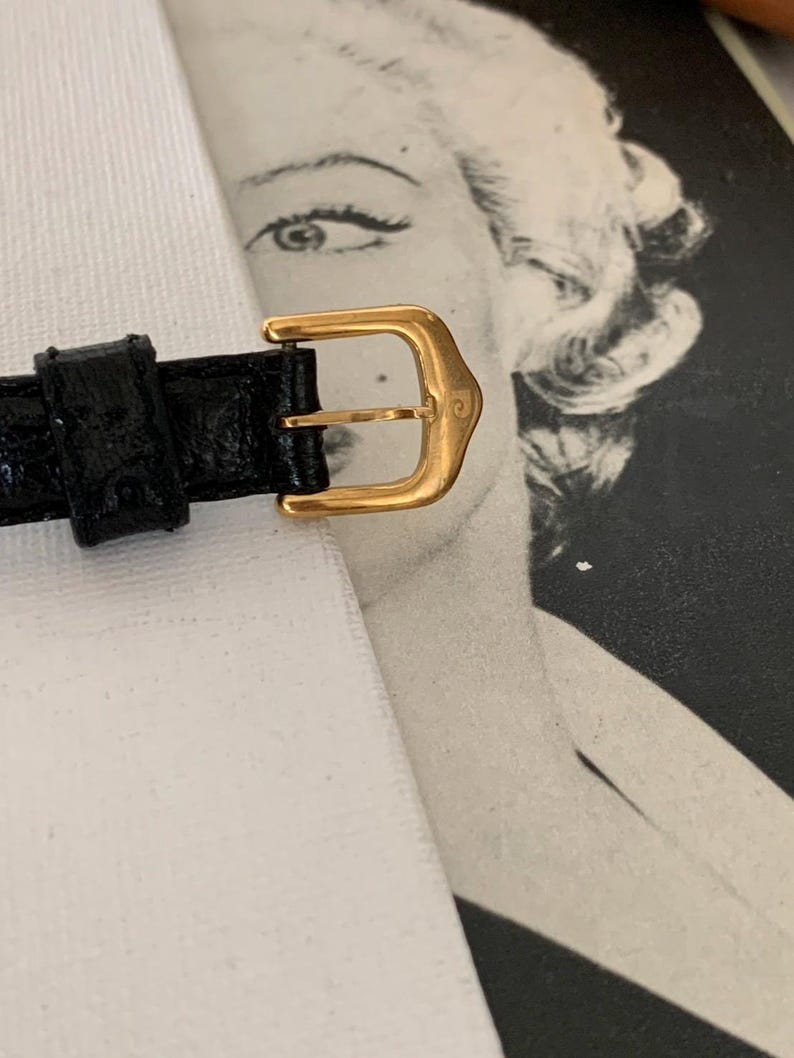May include: A gold-tone watch strap buckle with a black leather strap. The buckle has a simple design with a smooth finish.
