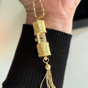 Puede incluir: Un collar dorado con un colgante de reloj. El reloj tiene una esfera rectangular con esfera negra y agujas doradas. El colgante está encerrado en un marco cilíndrico dorado texturizado, con una borla. La cadena también es dorada.