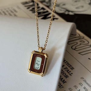 May include: A gold-coloured necklace with a rectangular pendant resembling a digital watch. The pendant has a dark red face displaying "ALBA" and the time "127". The chain is a fine link design.