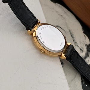 May include: A gold-tone wristwatch with a black leather band. The back of the watch case is silver and says "Stainless Steel Back" and "21322".