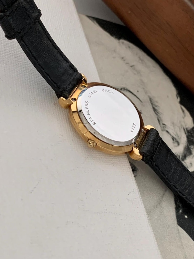 May include: A gold-tone wristwatch with a black leather band. The back of the watch case is silver and says "Stainless Steel Back" and "21322".