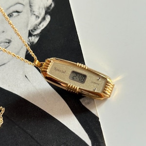 Puede incluir: Collar con colgante de reloj de cuarzo dorado. La esfera rectangular del reloj muestra la hora 2:48. La palabra "Special" está encima de la pantalla digital. El reloj está suspendido de una delicada cadena dorada. El fondo presenta una imagen en blanco y negro.
