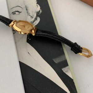 May include: A gold-toned wristwatch with a black leather band. The watch face is octagonal and has Roman numeral hour markers.