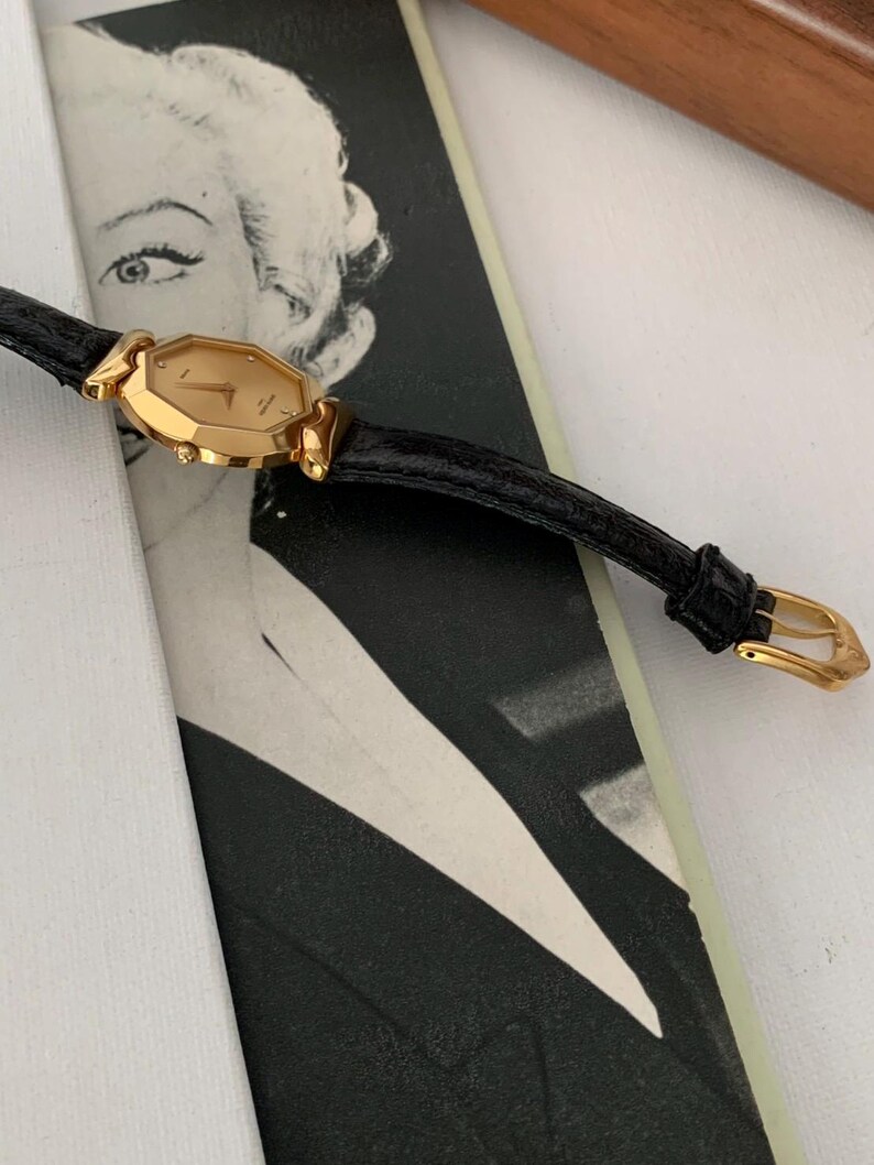 May include: A gold-toned wristwatch with a black leather band. The watch face is octagonal and has Roman numeral hour markers.