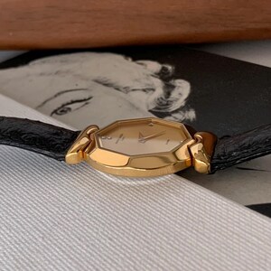 May include: A gold-tone wristwatch with a black leather band. The watch face is octagonal and has a white background with black Roman numeral hour markers. The watch has a date window at the 3 o'clock position.
