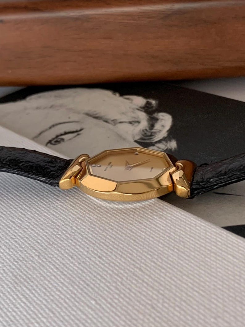 May include: A gold-tone wristwatch with a black leather band. The watch face is octagonal and has a white background with black Roman numeral hour markers. The watch has a date window at the 3 o'clock position.
