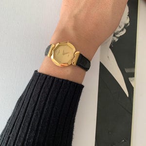 May include: A gold-toned watch with a black leather band. The watch face is octagonal and has a white dial with black Roman numeral hour markers.