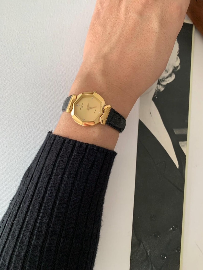 May include: A gold-toned watch with a black leather band. The watch face is octagonal and has a white dial with black Roman numeral hour markers.