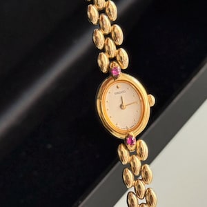 May include: A gold-toned Seiko wristwatch with a cream-colored face and gold hands. The watch features a gold band with oval links and two small pink gemstones. The watch is set against a dark background.